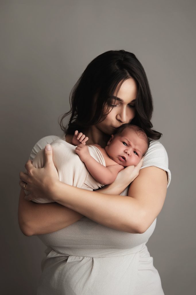 Jeune mère tenant son nouveau-né emmailloté contre elle, séance naissance en studio à Juziers par Rose Basile