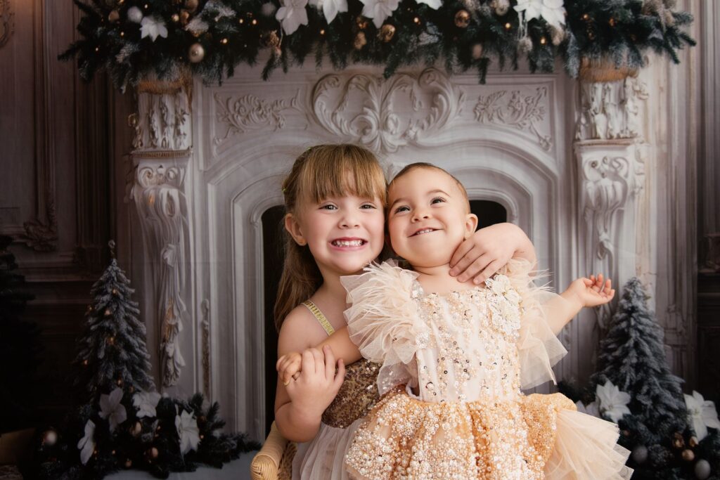 Deux sœurs souriantes se serrent l'une contre l'autre dans le décor de Noël à la cheminée du studio Rose Basile.