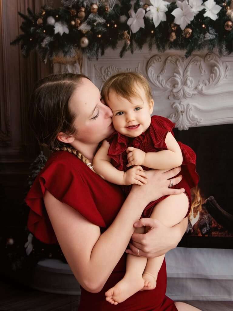 Une maman embrasse tendrement son bébé vêtu de rouge lors d'une séance photo de Noël devant une cheminée.