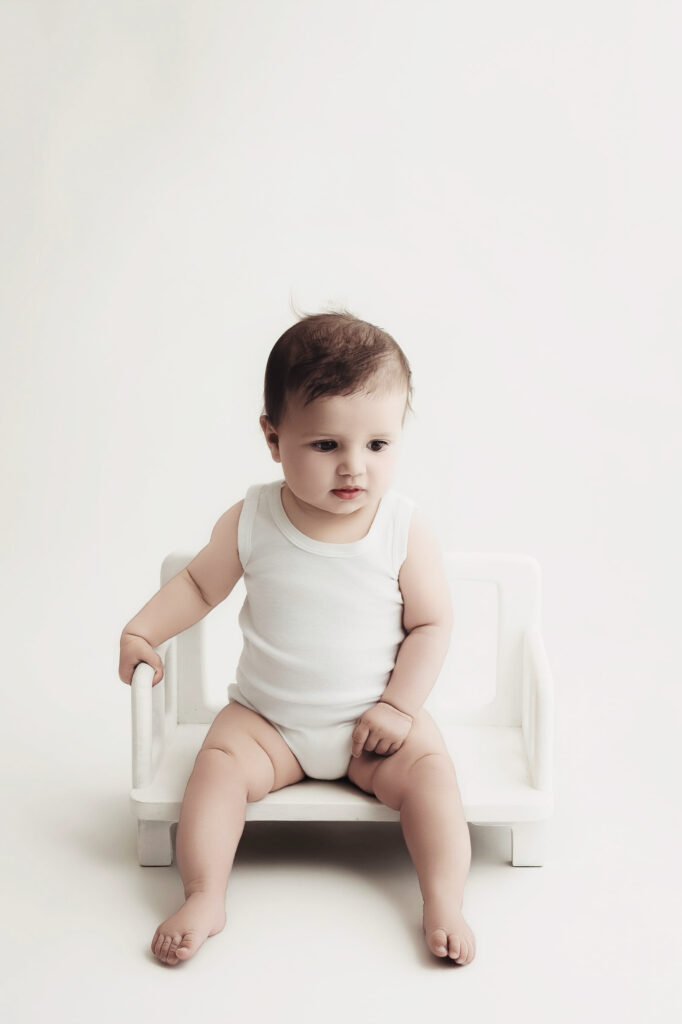 Bébé assis sur banc blanc, en body blanc, sur fond blanc