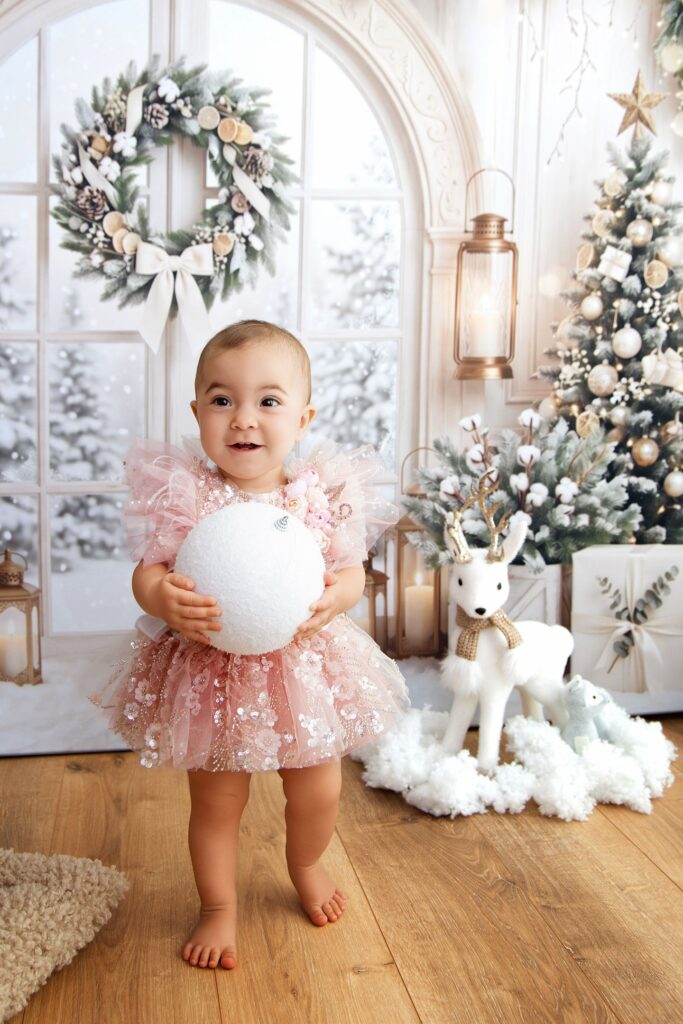 Bébé en robes de tulle rose, tenant une boule de neige devant une arche blanche et une fenêtre enneigée