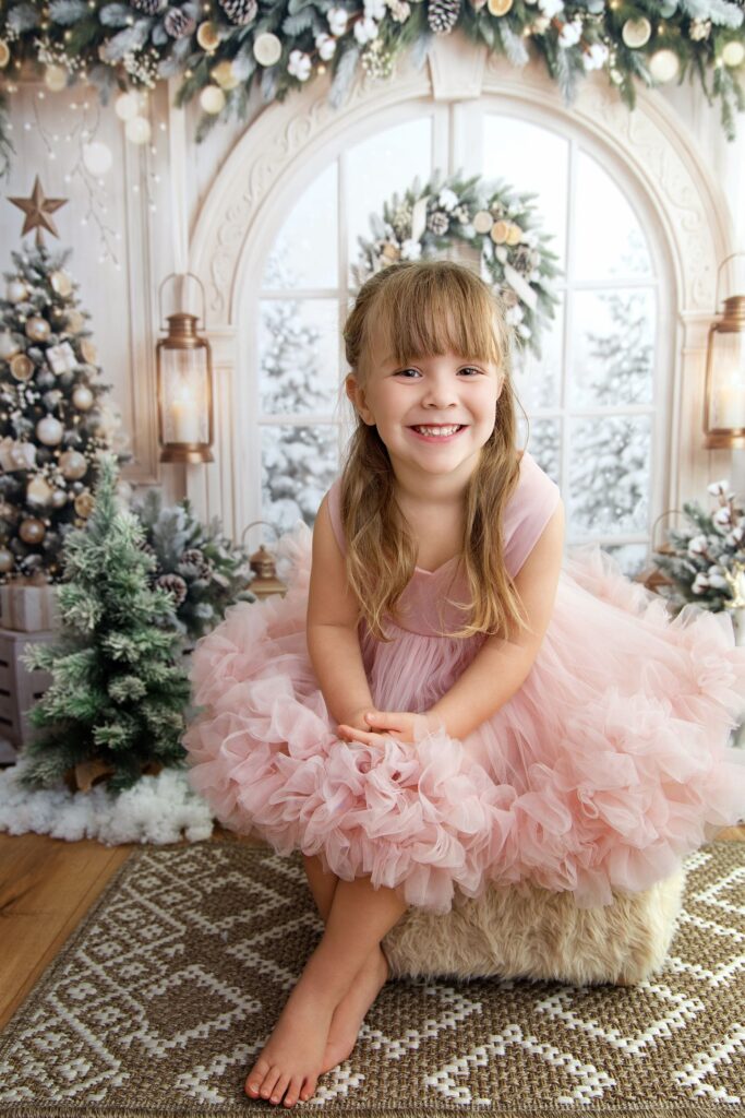 Fillette en robe de tulle rose, assise devant une arche blanche et une fenêtre enneigée