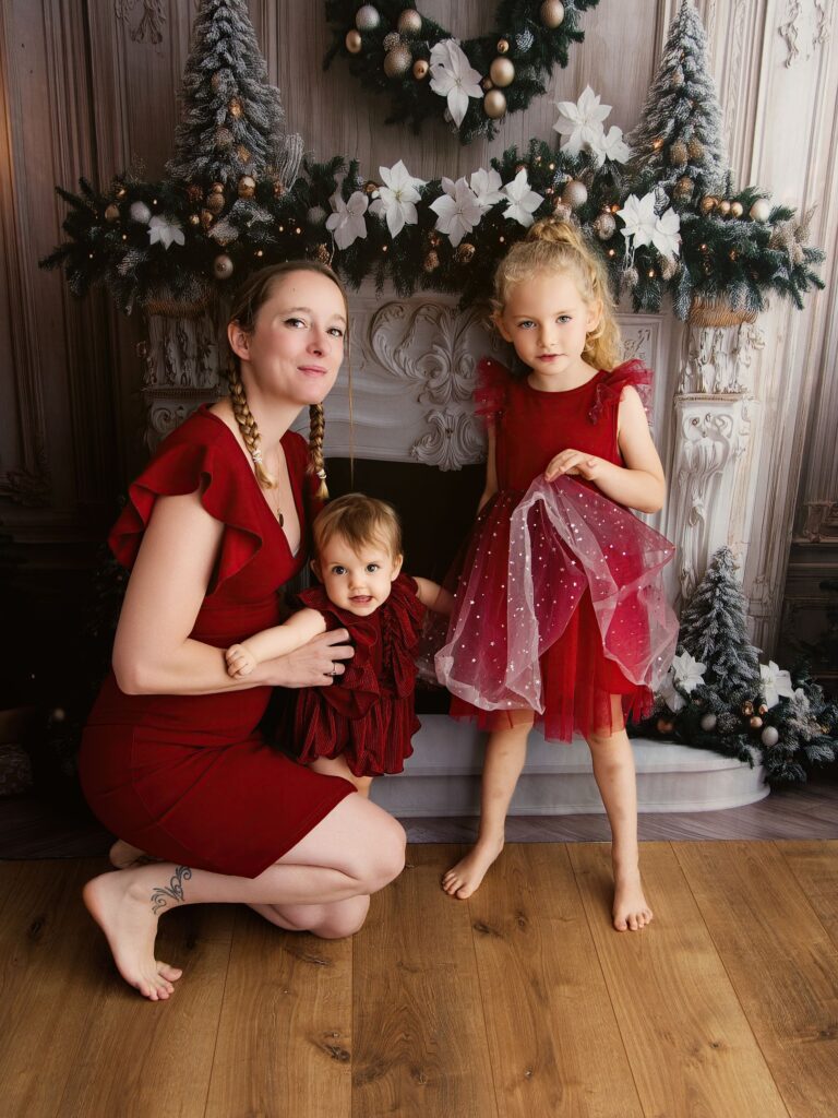 Une maman et ses deux filles en robes rouges réunies devant une cheminée décorée pour Noël au studio Rose Basile à Juziers.