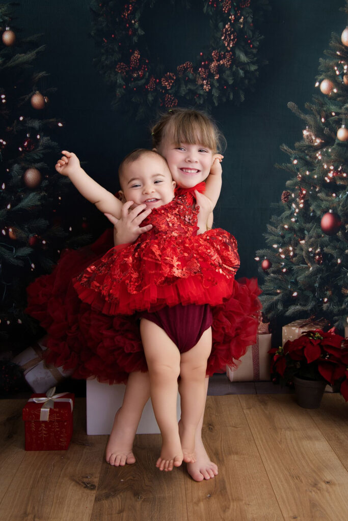 Deux sœurs en robes rouges, la grande serrant la petite dans ses bras, devant les sapins de Noël