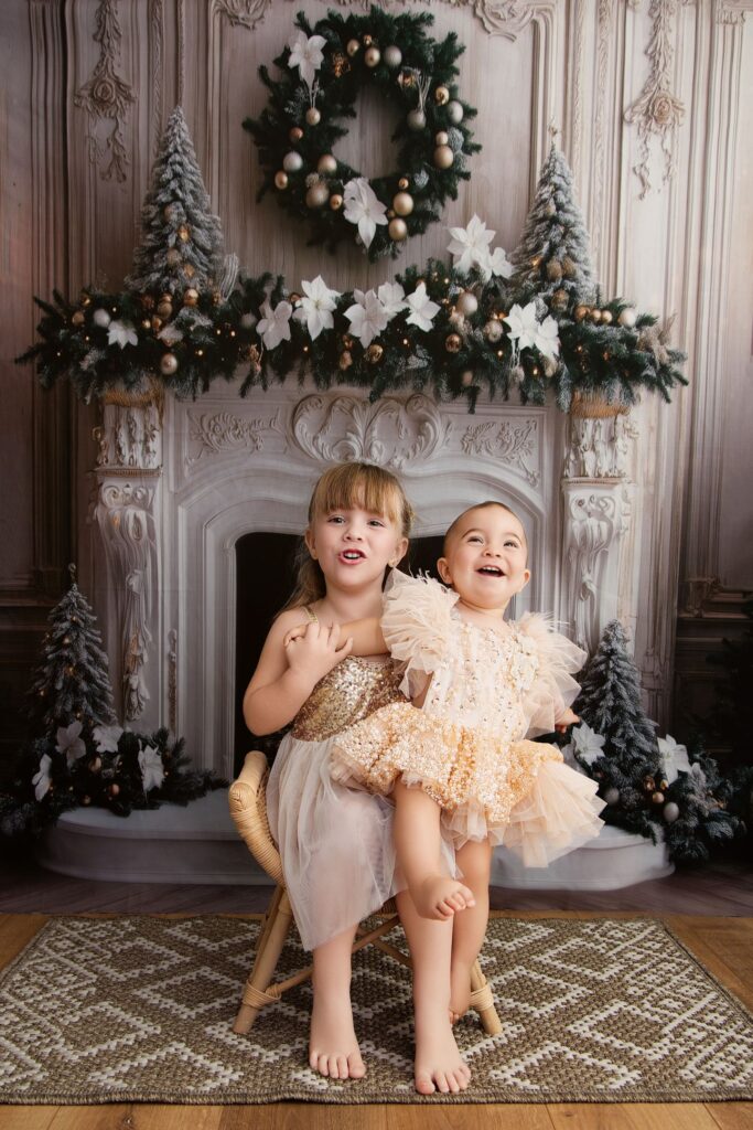 Deux sœurs en robes dorées qui rient devant une cheminée décorée pour Noël, séance Édition Noël au studio